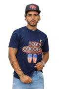 Man wearing a navy blue t-shirt with 'Soy Cocolo' text and graphics, standing against a white wall.